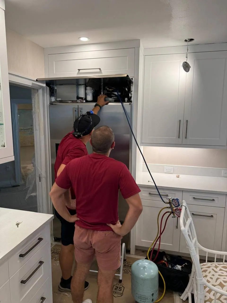 Two SunTech technicians in maroon shirts at a built-in stainless refrigerator, the top grille raised and a gauge hose running down to the floor.