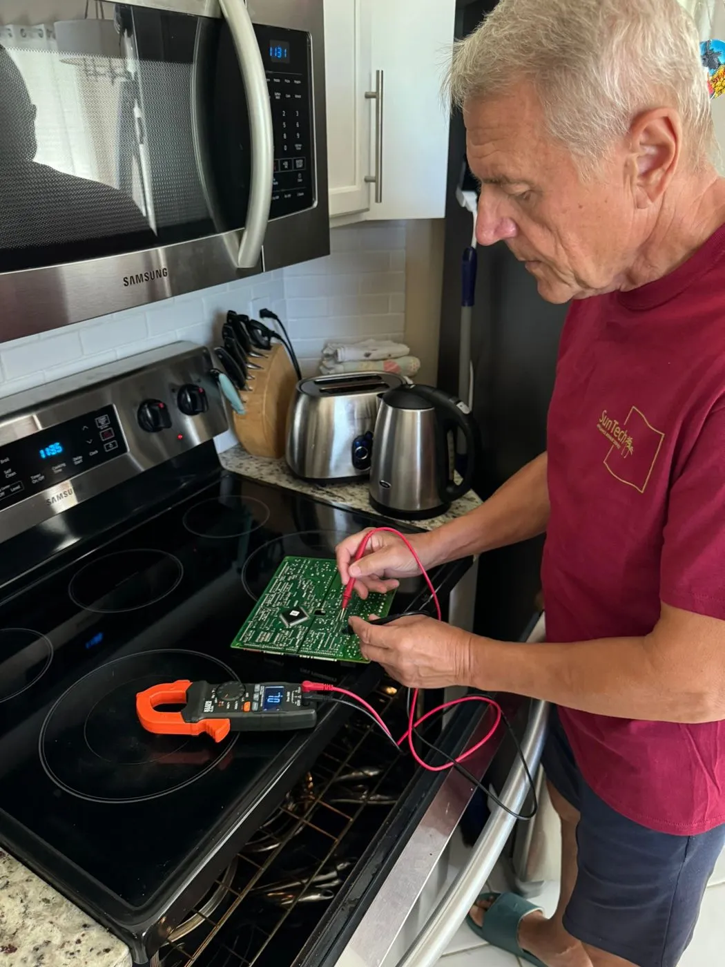 A technician tests a green control board with meter probes, the board laid on a glass cooktop.