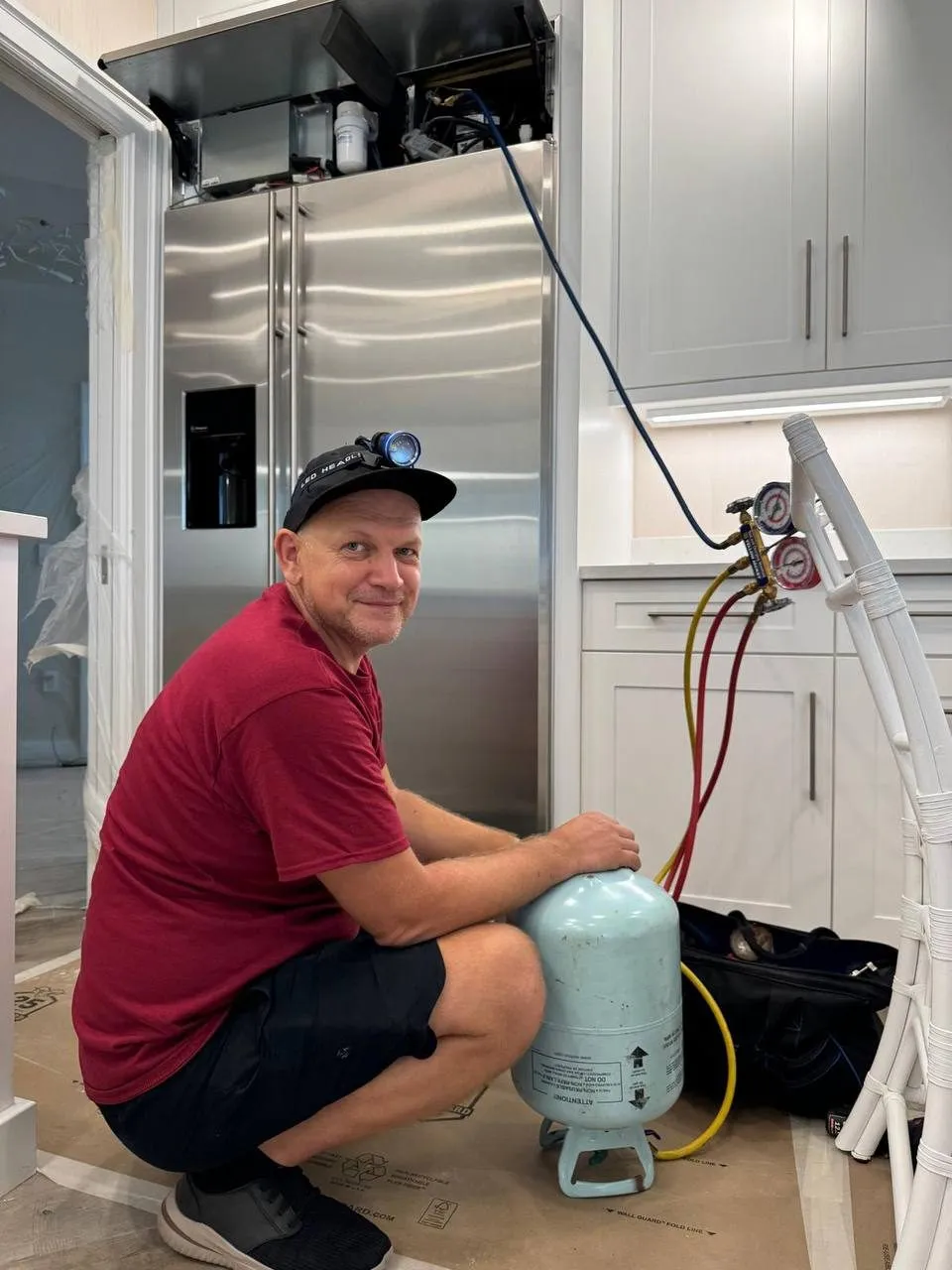 A technician in a headlamp crouches beside a refrigerant cylinder in front of a built-in refrigerator and smiles at the camera.