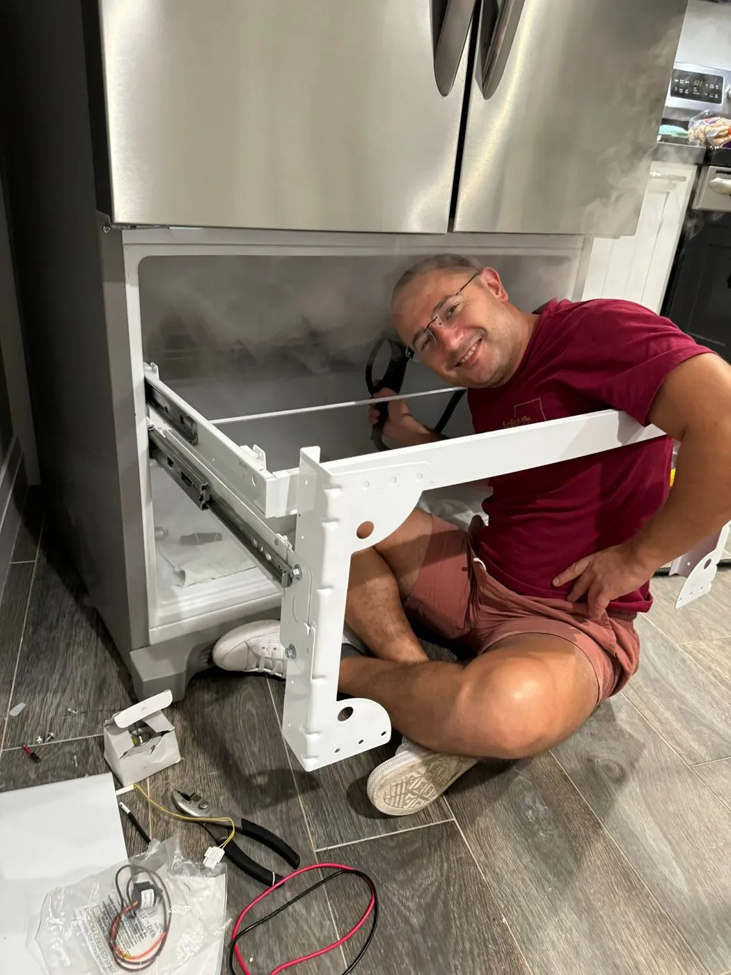 A SunTech technician sits on a kitchen floor with the drawer frame of a French-door refrigerator in his hands, smiling.