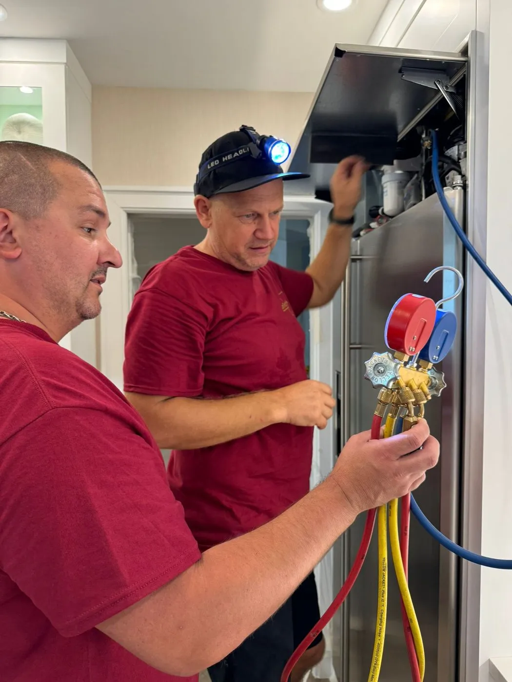 One technician holds a manifold gauge with red, blue and yellow hoses while the other, in a headlamp, reaches into the open top of the refrigerator.