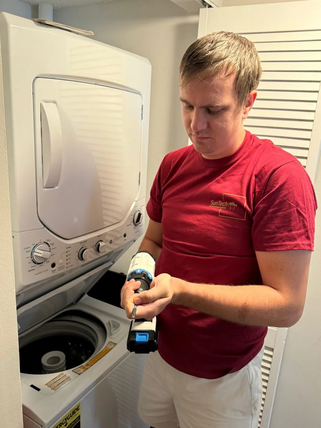A technician in a maroon SunTech shirt holds a drill beside a stacked washer and dryer in a laundry closet.